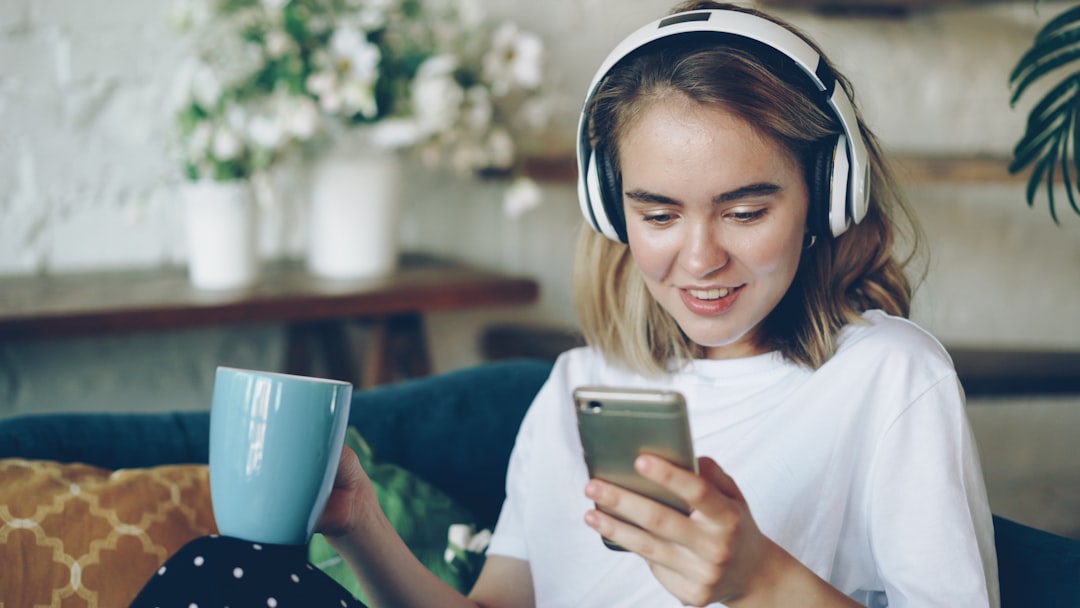Happy young lady is using smartphone chatting online and listening to music with wireless headphones sitting on sofa in modern flat. Technology, people and joy concept.