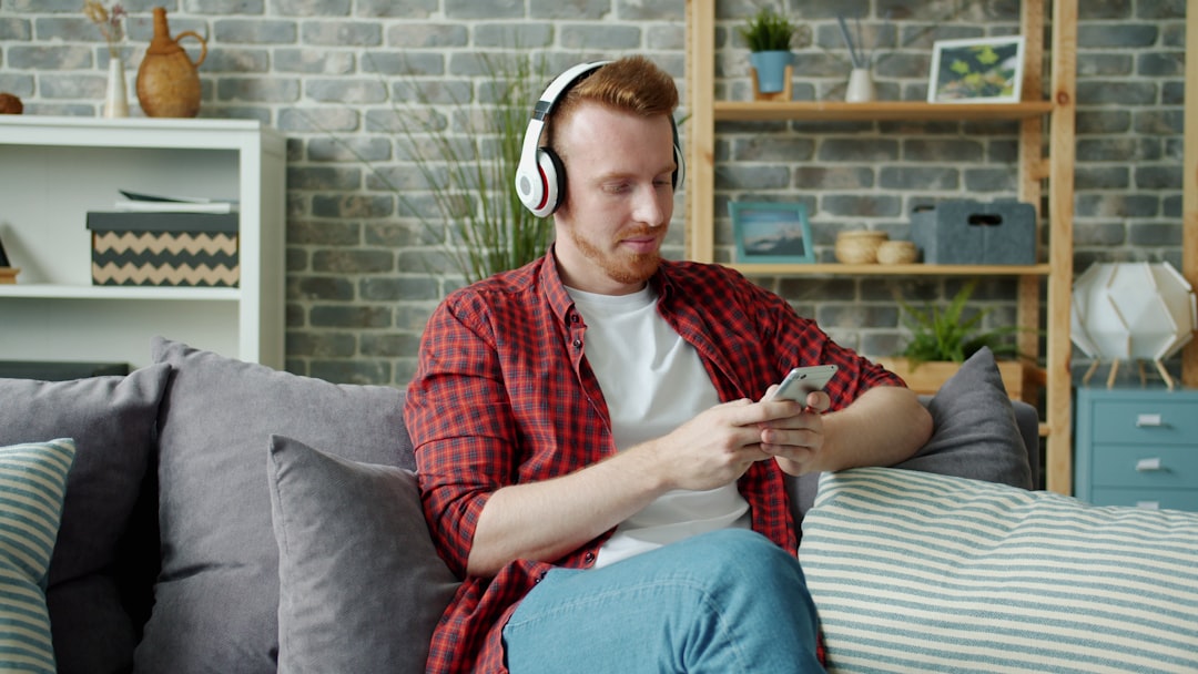 joyful guy in wireless headphones listening to music and using smart phone relaxing inside beautiful cozy apartment. Youth and devices concept.
