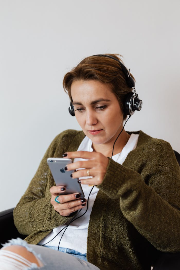 Content female wearing jeans and warm jacket sitting against white wall and browsing contemporary cellphone while enjoying songs in headphones