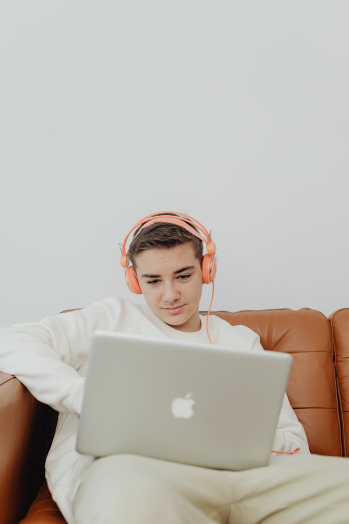 Teenager wearing headphones while using a laptop indoors, relaxing on a couch.